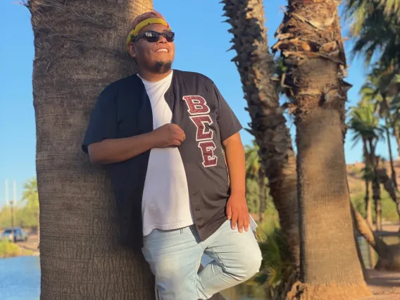 Portrait of a male presenting individual leaning against a palm tree