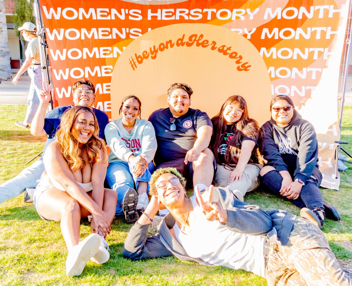 A group of students and staff sitting on the grass in front of a Women's Herstory Month backdrop