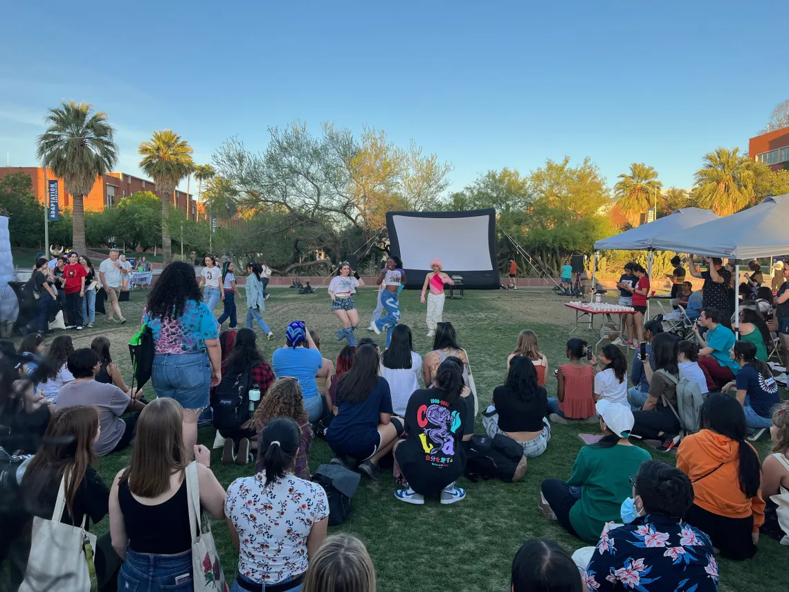 A group of students sitting outside on the grass watching students perform a dance routine