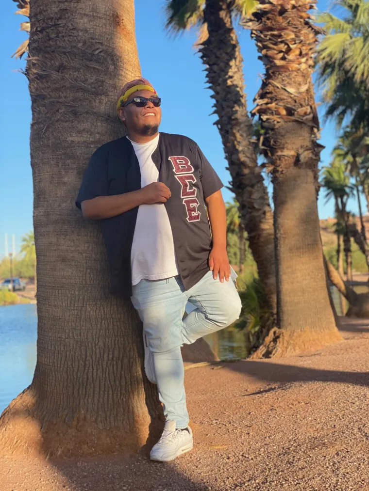 Portrait of a male presenting individual leaning against a palm tree